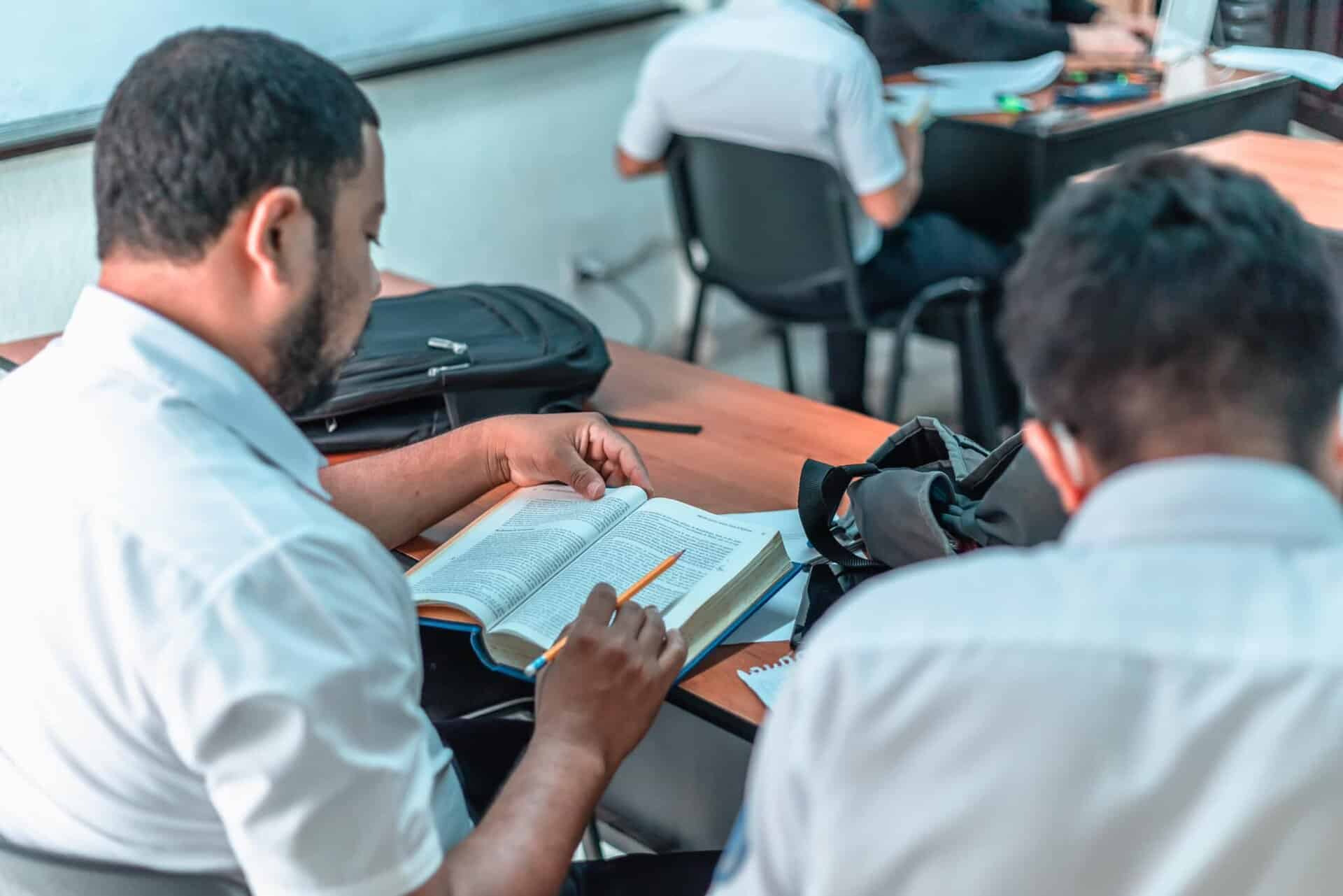Two men sit at a desk in a classroom, one reading a textbook and holding a pencil. Other students are seated in the background.
