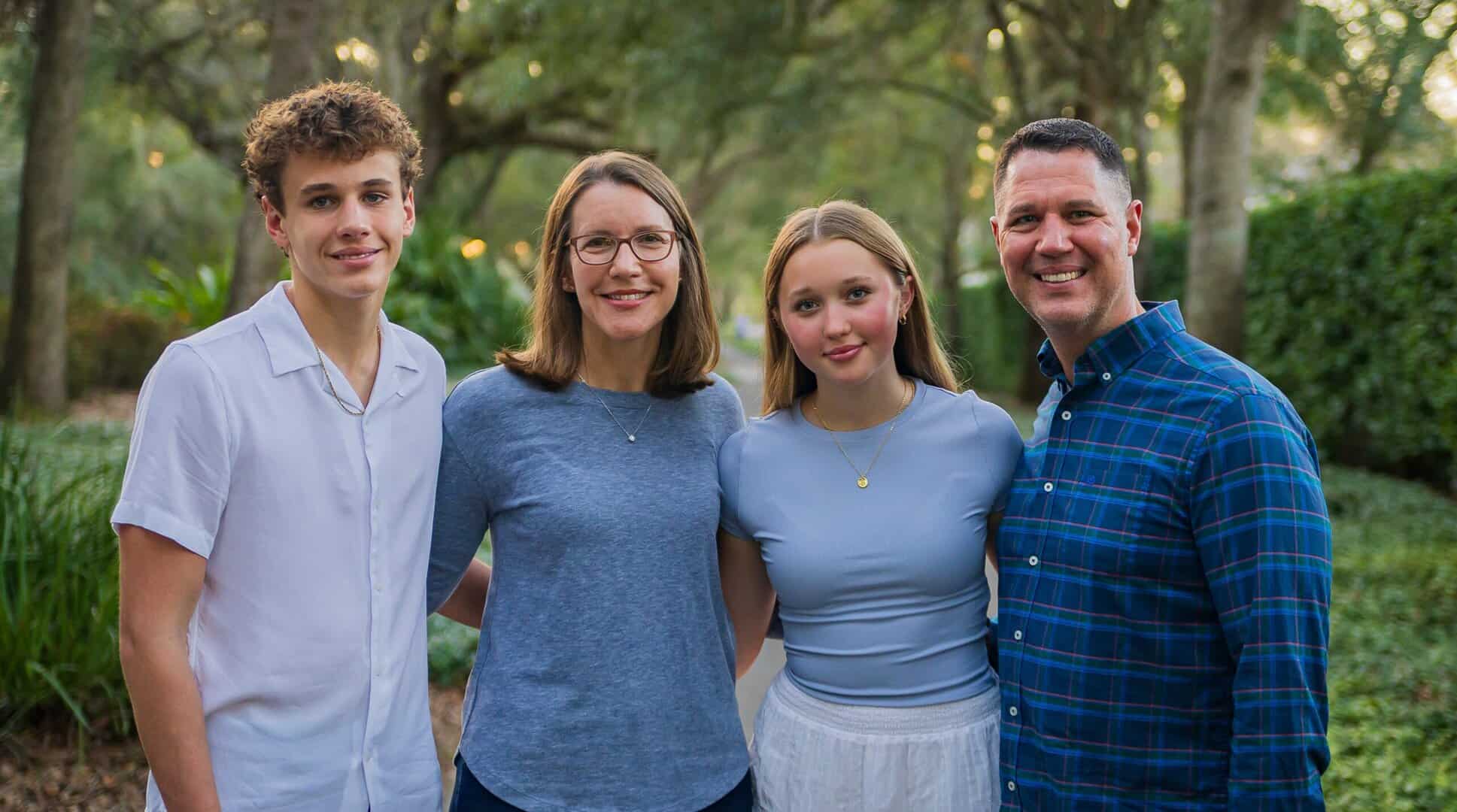 A group of four people, two adults and two teenagers, stand outdoors in a park-like setting, smiling at the camera.