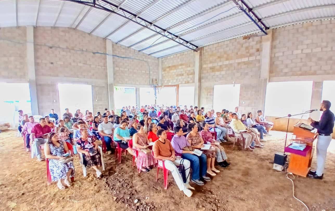 A group of people seated on plastic chairs in a large, unfinished building, listening to a speaker at a podium in front.