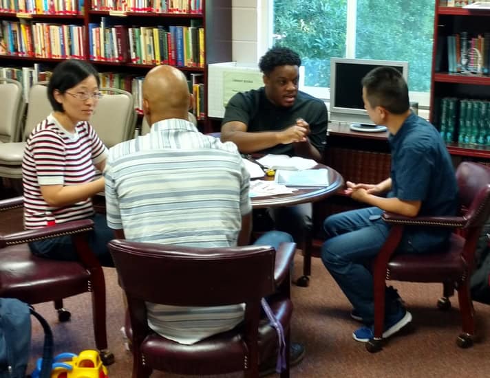 Four people sit around a table in a library, engaged in discussion. Bookshelves and a computer are visible in the background.