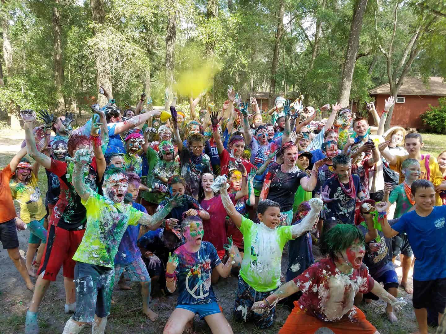 A large group of people outdoors covered in colorful paint, cheerfully raising their hands, with trees and a cabin in the background.