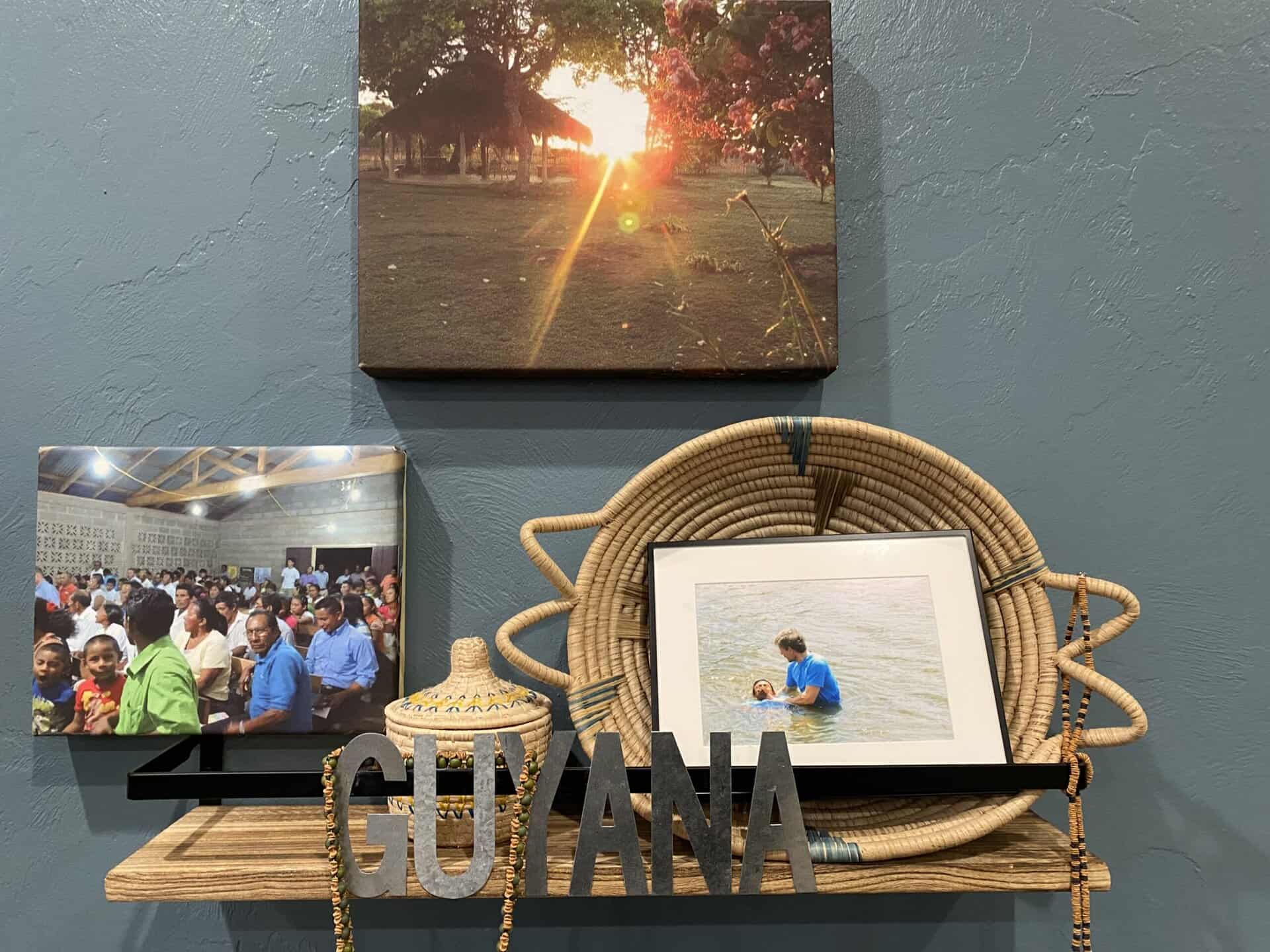 A shelf display with "GUYANA" letters, a woven basket, a photo of a child in water, a group photo, and a sunset picture.