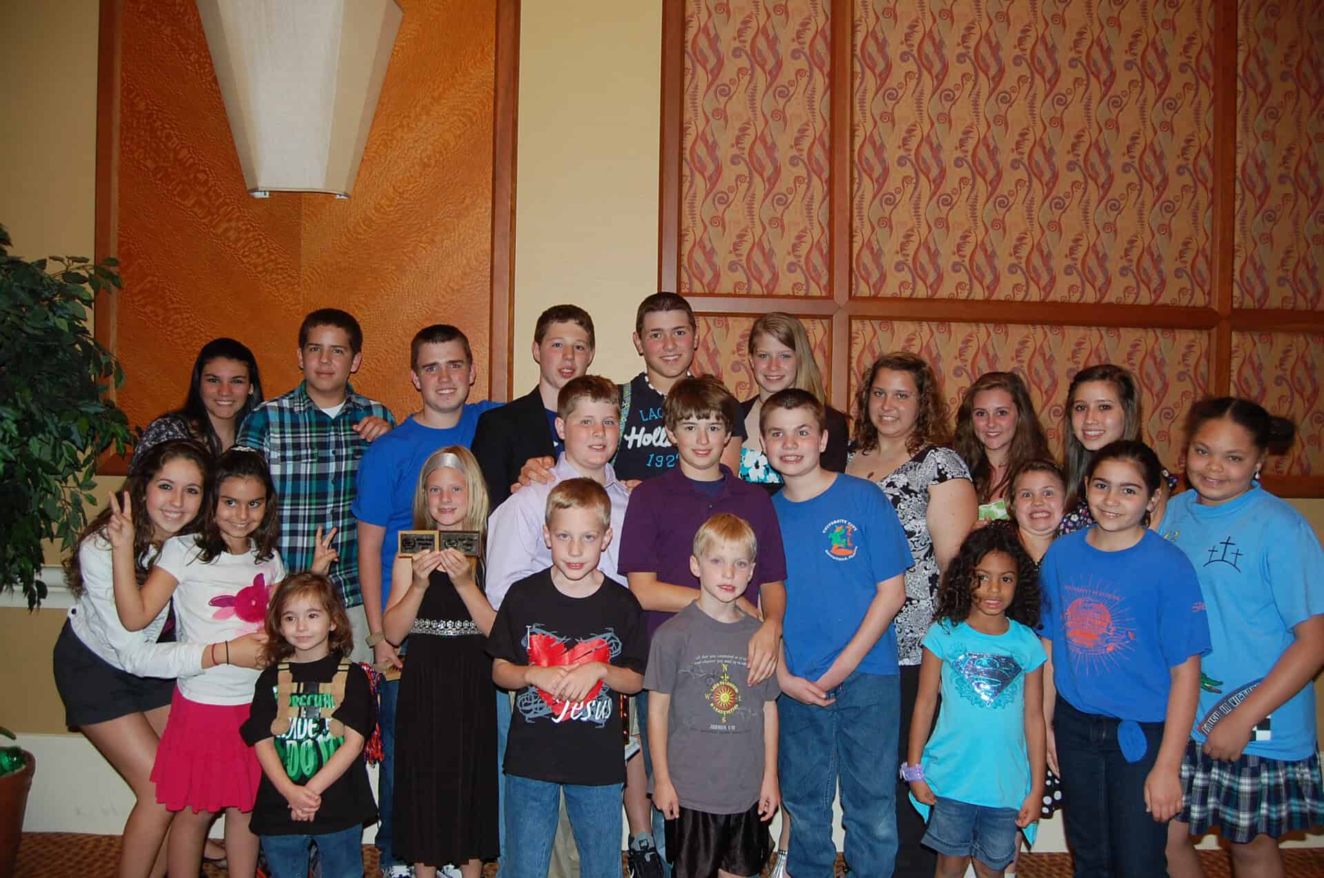 A group of people of varying ages, including adults and children, are standing together indoors, smiling at the camera.