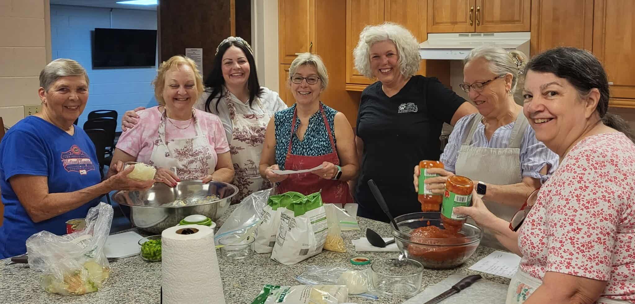 Seven women stand and smile together in a kitchen while preparing food, with ingredients, mixing bowls, and utensils spread on the counter in front of them.