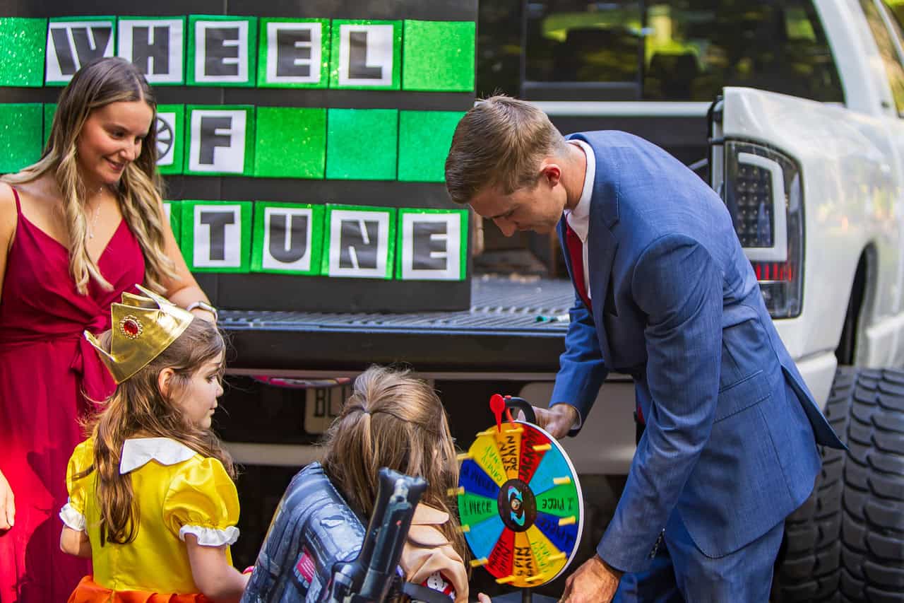 A man in a suit helps children spin a colorful wheel labeled "Trick or Treat" beside a "Wheel of Fortune" sign. A woman in a red dress observes. They're at the back of a pickup truck.