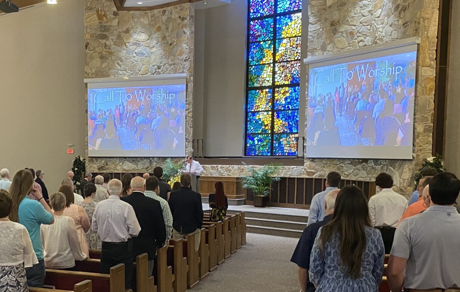 A congregation stands in a church, facing a stage with projected screens showing "Call to Worship." Stained glass windows are visible in the background.