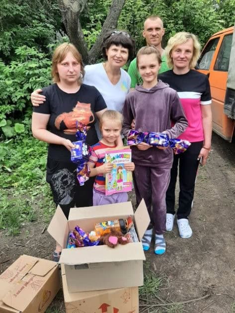 A group of six people, including three adults and three children, stand outdoors beside boxes filled with wrapped gifts and a magazine. Trees and an orange vehicle are in the background.