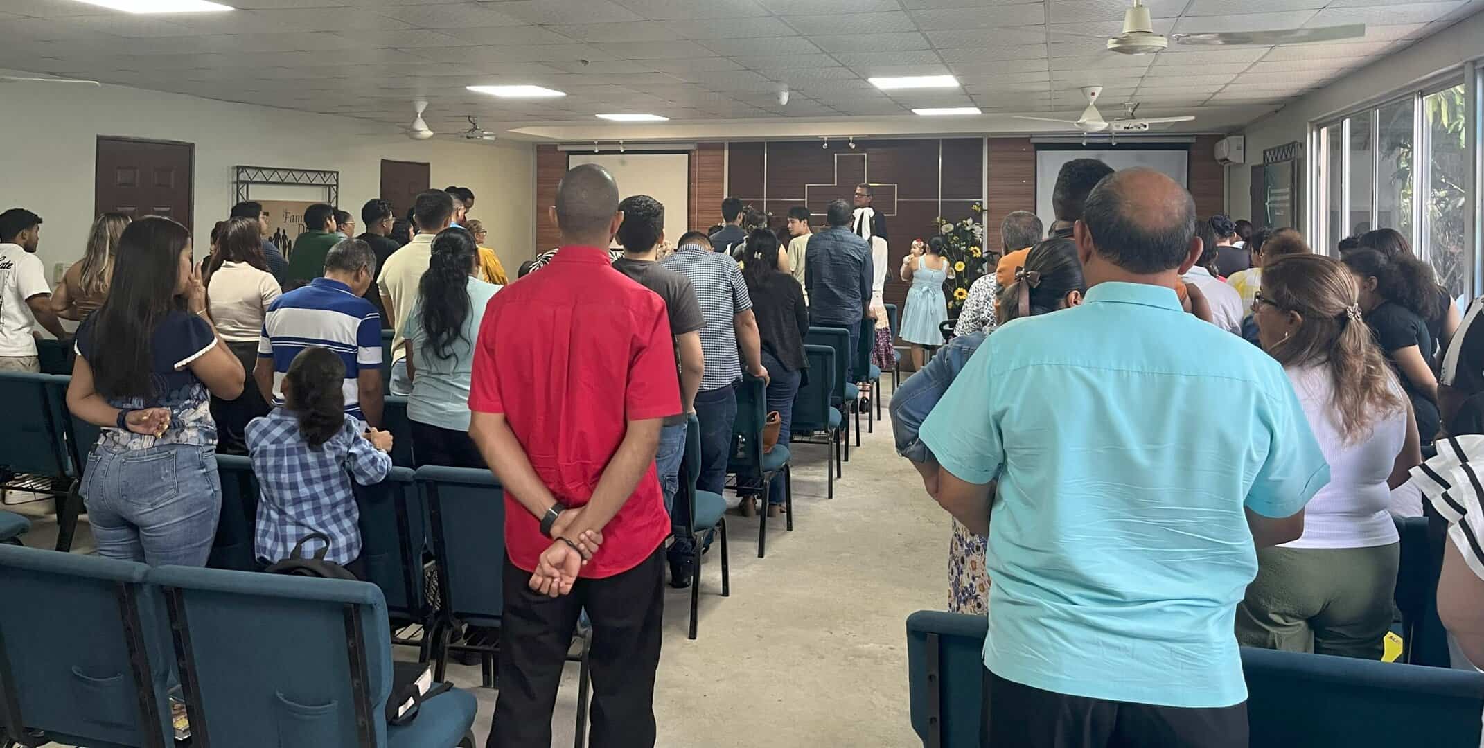 A group of people stand inside a church, facing the front where a speaker is standing at a podium. Some attendees have their heads bowed.