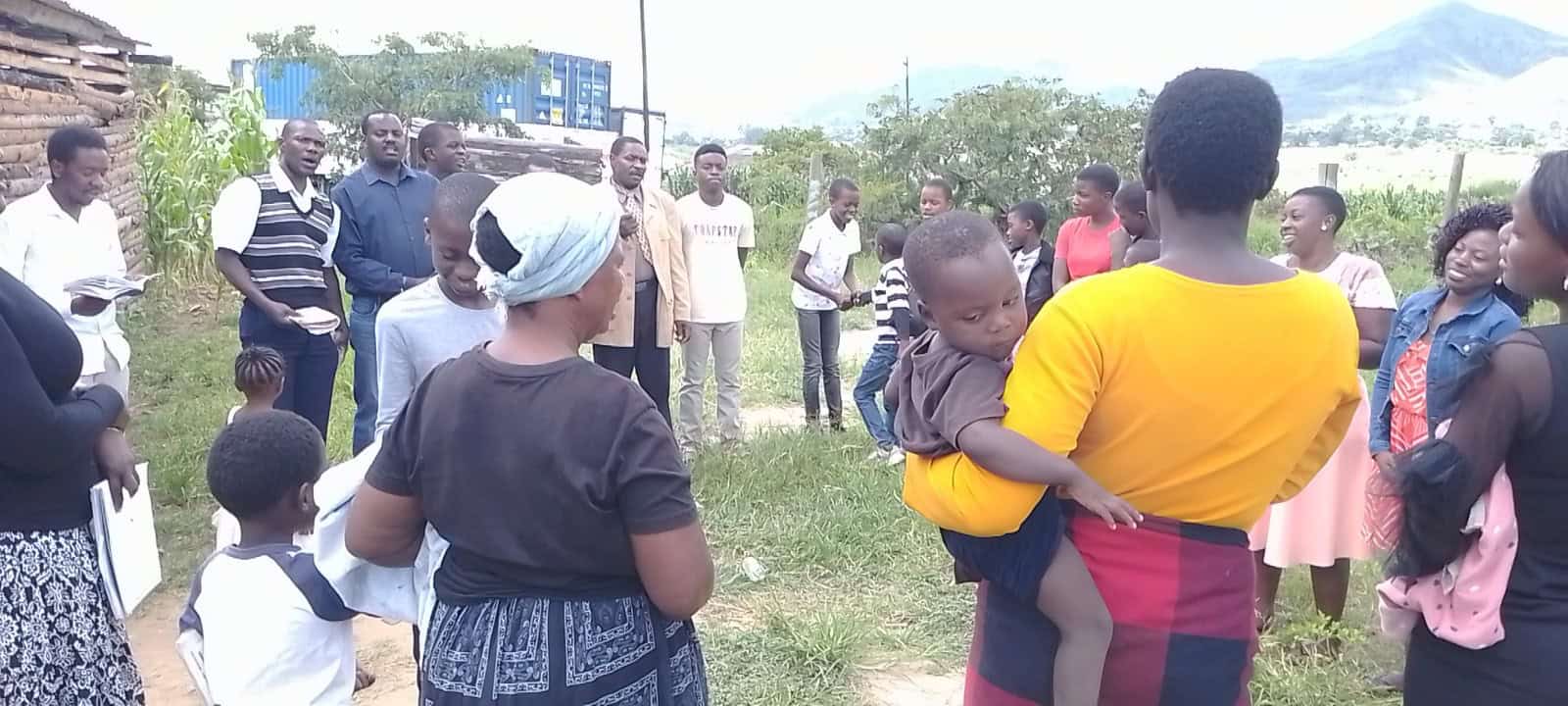 A group of people stand outdoors in a circle on grass. A woman in a yellow shirt holds a child. Some individuals hold papers, and a mountain is visible in the background.
